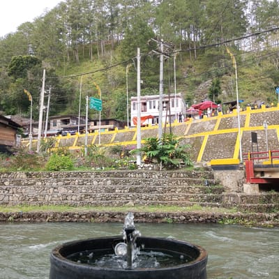 In the city of Blang Bebangka, Indonesia, a river flows in the foreground, with a black cylindrical object containing a small fountain in the center. Behind the river, a stone wall with a yellow geometric pattern rises up a slope. A few people are visible on top of this wall, near buildings and red tents. Further up the slope, a dense forest of tall, thin pine trees covers the hillside. Power lines crisscross the scene, supported by utility poles. A road sign points towards "Kota Bandara Rembula-Bireuen" and "Luar Garut-Luar Ail Lampan".