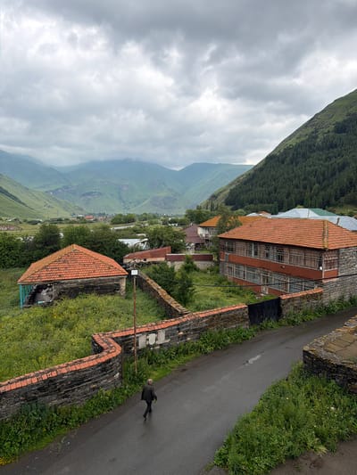 A person walks on a wet asphalt road in the village of Sno, Georgia, on a cloudy day. The road is lined with a stone and brick wall covered in greenery. Behind the wall, there are several buildings with orange tiled roofs, some appearing old and weathered. Further in the background, more houses and trees are visible, nestled in a valley. The landscape is dominated by large, verdant mountains under a heavy, overcast sky. The overall scene suggests a rural, mountainous village setting.