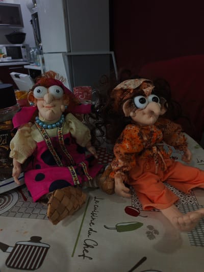 Two handmade dolls, likely trolls, are positioned on a table covered with a tablecloth. The doll on the left has bright pink polka dot overalls over a cream-colored blouse, and a red bandana. It wears a blue beaded necklace and has large, googly eyes. The doll on the right wears an orange floral-patterned blouse and orange pants. It also has large, googly eyes and its hair appears to be brown. The tablecloth has a "La Cuisine du Chef" inscription and images of kitchenware like pots and peppers. In the background, out of focus, are kitchen appliances. The lighting suggests an indoor setting, likely during the day. The dolls appear to be posed for a photograph.