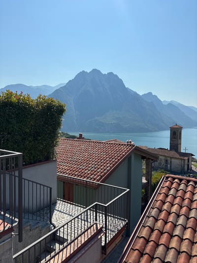 A clear, bright day captures a view from a village in Zorzino, Italy, looking out towards a large, calm body of water and distant, mist-shrouded mountains. In the foreground, several red-tiled roofs are visible, one of which has a chimney. A well-maintained green hedge is situated on a lower level, with a metal railing and stairs leading down. To the right, a church tower with a clock face stands next to a stone building, complete with a cross on its roof. The water is a serene turquoise, reflecting the clear blue sky and the surrounding mountains, which display varying shades of blue and green due to atmospheric perspective. The village buildings are constructed with traditional materials, featuring terracotta roof tiles and stone elements. The overall impression is one of peaceful, picturesque Italian village life against a dramatic natural backdrop.