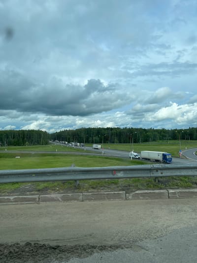 The image shows a highway scene near Starokurovo, Russia. The view is from an elevated position, possibly a bridge, looking along a multi-lane road with a median. A line of commercial trucks is visible on the highway. Beyond the road is a grassy strip and a dense line of trees. A cloudy sky fills the upper portion of the image. A metal safety barrier is in the foreground. A roundabout is ahead of the line of trucks.