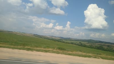 The image depicts a panoramic view of a landscape with rolling green hills under a partly cloudy sky. In the foreground, a dirt road with a white line runs diagonally across the frame. The hills in the midground are covered in green vegetation, with patches of lighter brown suggesting dry grass or tilled fields. Scattered trees are visible in the distance, and the terrain becomes more rugged and mountainous towards the horizon. The sky is a bright blue, dotted with white cumulus clouds, some of which are large and dramatic. The overall impression is of a rural or natural setting. This image appears to be taken from a moving vehicle, as evidenced by the slight motion blur in the foreground. The location is identified as Zmeinogorsk, Russia.