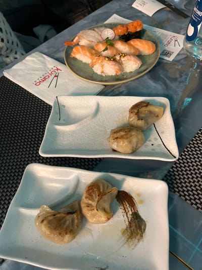 An overhead shot captures a dining table set with various Japanese-style dishes and tableware. The scene depicts a meal being served or consumed in what appears to be a restaurant setting.

On the left, an oval, grey-green ceramic plate holds an assortment of nigiri sushi, including salmon and shrimp. Some pieces are topped with small red fish or shrimp, and the plate is garnished with a thin slice of radish and a small herb sprig. To the right, two white, rectangular plates, each holding two pan-fried or steamed dumplings (gyoza), are visible. One of these plates features an artistic brown brushstroke design, possibly made with a sauce or oil.

The table features dark, patterned placemats and a blue-grey tabletop. A clear glass bottle with a blue label, partially obscuring the word "ANTONIO" and "SPECIALE," is seen in the upper left. A white paper napkin with the distinct red logo and text "kūrama RESTAURANT" is positioned near the sushi plate. A white receipt or bill is also partially visible on the left. The overall impression is an indoor evening meal at a Japanese or Asian fusion restaurant, possibly in Basiglio, Italy, as indicated by the context.