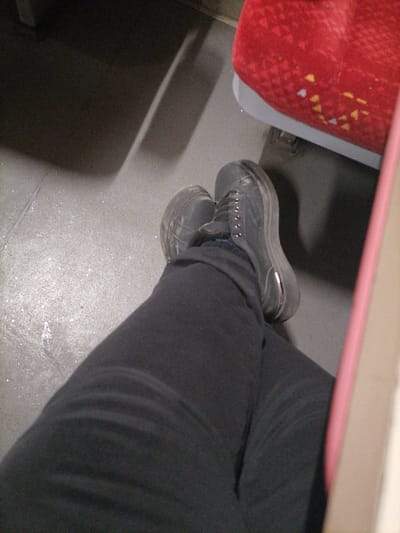 The scene is set inside a public transport vehicle in Tompsan, Bulgaria. The image, captured from a low, seated perspective, features the lower extremities of an individual. The subject wears dark-colored trousers and dark grey or black laced shoes with prominent light-colored soles, exhibiting signs of wear. A portion of a blue sock is visible above the right shoe. The feet rest on a light grey, speckled floor surface. To the upper right, a segment of a red upholstered seat is visible, characterized by small, irregular yellow and white patterns and a light grey lower trim. Elongated shadows are cast across the floor from unseen objects. A vertical, narrow pinkish-beige structure partially borders the right edge of the frame.