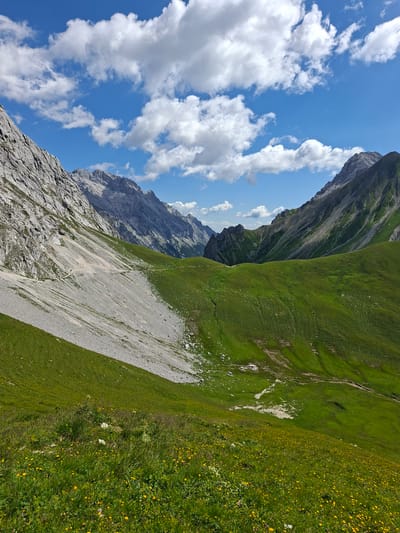 The image depicts a mountainous landscape with lush green hills and rocky slopes under a blue sky dotted with white clouds. In the foreground, a meadow is covered with vibrant green grass and numerous small yellow wildflowers. A path winds its way up a scree slope on the left side of the image. Further into the distance, more mountain ranges are visible, some with sparse vegetation and others appearing rocky and bare. The scene conveys a sense of natural beauty and remoteness. This location is identified as being in the municipality of Mieming, Austria.