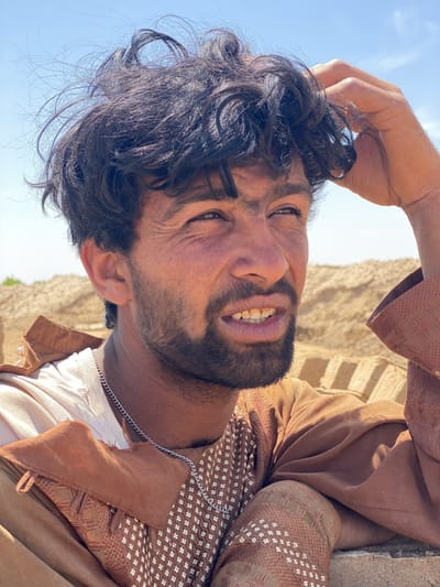 A medium shot shows a man in Deh-E Bagh, Afghanistan. He has dark curly hair and a beard. He wears a white t-shirt under a brown embroidered garment and a silver chain around his neck. His right hand is raised to his head. The background includes a blue sky and a tan structure, possibly made of bricks.
