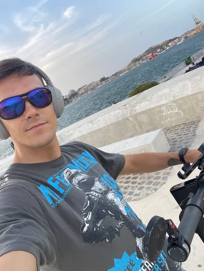 This is a close-up, selfie-style photograph of a man with short brown hair, wearing large grey over-ear headphones and mirrored sunglasses. He is wearing a dark grey t-shirt with a graphic print featuring blue lettering and an image of a person on a motorbike. The man is positioned in the foreground, with his face turned towards the camera.
In the background, there is a large body of blue water, likely the Mediterranean Sea. A shoreline with buildings and a hill is visible in the distance. The man appears to be standing on a paved walkway or promenade, made of cobblestones, with a concrete barrier to his right. The scene is set in the city of Cartagena, Spain, specifically along its waterfront. The lighting suggests it is daytime, possibly late afternoon, with soft sunlight casting a gentle glow. The overall impression is one of a casual outdoor setting, likely a leisure activity.