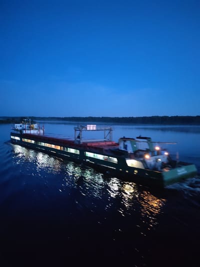 A long, illuminated cargo barge moves on a body of water under a darkening sky. The barge has multiple light sources visible along its length. Reflections of these lights streak across the water's surface. A line of trees is visible on the horizon. The city of Gari, Russia, is not explicitly visible in the image, but the presence of such a large barge suggests the location is near a major waterway in the region.