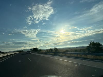 The image shows a view from a car driving on a highway, likely in or near Vinarovo, Bulgaria, given the context. The highway has multiple lanes and is paved with asphalt. A silver car is visible in the distance, traveling in the same direction. The road is bordered by a metal guardrail and a grassy embankment with scattered trees and bushes. The landscape beyond the embankment appears to be rural, with fields and some distant buildings or structures. The sky is filled with a mix of white and gray clouds, with the sun shining brightly through a gap, casting a warm, golden light. The overall atmosphere suggests a clear, bright day, possibly late afternoon or early morning.