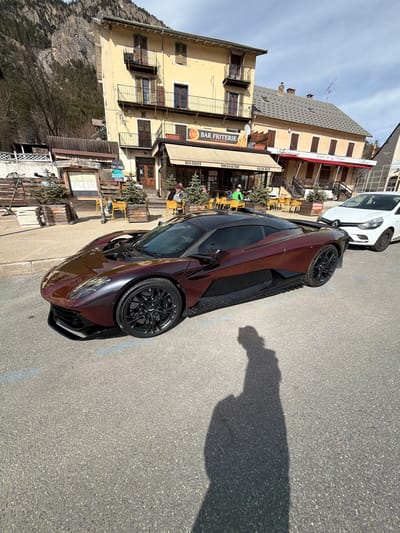 This image depicts a street scene in Briançon, France, specifically on or near Rue de la Digne, with the prominent yellow building identified as "BAR FRITERIE" and "BISTROT / BRASSERIE / GLACIER" at street level.

The foreground is dominated by a metallic maroon/reddish-brown high-performance sports car, likely an Aston Martin Valkyrie-like model, featuring a black roof and black multi-spoke wheels. It is parked on asphalt pavement, angled slightly left, with its front left quarter visible. A distinct human shadow, likely that of the photographer holding a device, is cast onto the pavement in the bottom right corner.

In the mid-ground, to the left of the yellow building, is an outdoor seating area associated with "Café Champ de Mars," furnished with wooden planters containing small fir trees and wooden benches. Several individuals are visible seated at yellow tables under a beige awning in front of the "BAR FRITERIE," engaging in casual outdoor dining or conversation; one person wears a bright green top. Adjacent to the yellow building on the right, another structure with a red awning houses "Restaurant" and "Café de la Digne." Further down the street, behind the sports car, a white Renault Clio is parked. The distant background reveals steep, rocky mountainsides with sparse vegetation and remnants of snow or ice, characteristic of an alpine environment. The scene is illuminated by strong daylight under a clear sky, casting pronounced shadows.