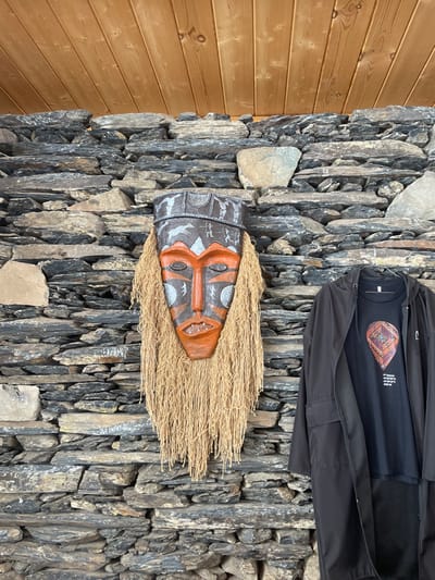 The scene depicts an interior space in Juta, Georgia. A prominent, elongated wooden mask is affixed centrally to a wall constructed of irregular, dark grey and brown dry-stacked stones. The mask features an orange face with grey and white painted patterns, dark outlines, and a dense, light-brown fibrous beard extending from its sides and bottom. Above the stone wall, a light-brown wooden plank ceiling is visible. To the right of the mask, a black hooded jacket is suspended, partially obscuring a black t-shirt underneath. The t-shirt displays a colorful, patterned, tear-drop shaped graphic, beneath which white text includes geographic coordinates and the explicit label "Juta, Georgia". No individuals are present in the scene.