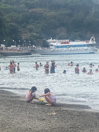 The image displays a bustling coastal scene in Sestri Levante, Italy, specifically within a bay featuring a pebbled beach and a pier.
In the foreground, two young girls are positioned on a dark, coarse-grained beach near the water's edge. The girl on the left wears a pink swimsuit and is kneeling, looking down at a yellow bucket. The girl on the right, wearing a lavender swimsuit with white detailing on the back, is also kneeling and appears to be interacting with sand or small objects near the bucket and a green shovel. A pair of light-colored flip-flops and a small green object are visible on the sand nearby.
The midground is dominated by calm, gray-toned bay water populated by numerous individuals wading or standing. These people, a mix of adults and children, are dressed in various swimsuits. Several yellow spherical buoys are scattered across the water's surface.
In the background, a large, multi-deck passenger boat with a white hull, a horizontal blue stripe, and red safety railings/life rafts is positioned near a stone pier. A considerable crowd of people is visible on the pier, some clustered near the boat's gangplank, indicating boarding or disembarking activity. White spherical streetlights line the pier. Behind the pier, a steep, densely forested hillside rises abruptly, showing hints of structures or pathways amidst the trees. The sky is overcast or hazy, creating diffused lighting across the scene.