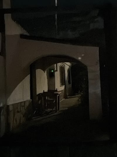 The image depicts a dimly lit archway leading into an outdoor area. The archway itself is constructed of textured white walls. To the left of the arch, a portion of a building wall is visible, with a lower section covered in rough-hewn stones. Inside the archway, a small table and chairs are positioned, appearing to be made of dark wood. Further back, an open doorway or window is visible, with a faint green light emanating from within. The overall environment suggests an exterior space, possibly a courtyard or alleyway, at night. The location is identified as Montgó, Spain.