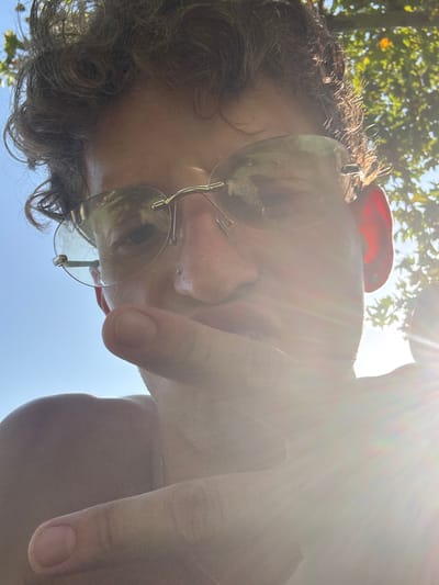 A person is taking a selfie outdoors, the sun is bright and shining directly into the camera lens, creating lens flare. The person has curly brown hair and is wearing rimless sunglasses. Their right hand is in the foreground, partially obscuring their face. In the background, there are green leaves and blue sky, suggesting an outdoor setting with trees. The sun's glare makes it difficult to discern fine details of the person's face or surroundings. The location is identified as Villa del Prado, Spain.