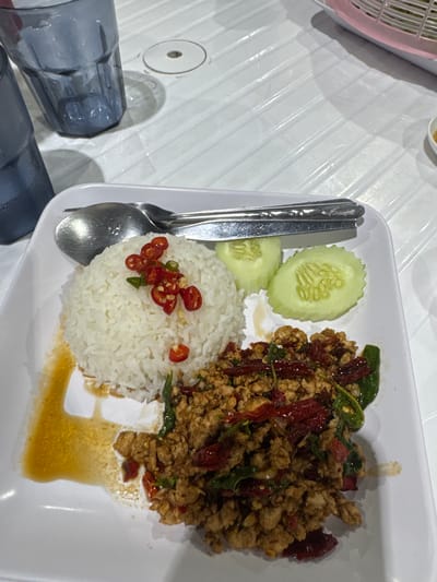 A plate of Pad Kra Pao, Thai basil chicken stir-fry, rests on a white table in Na Kluea, Thailand. The dish consists of stir-fried ground chicken mixed with basil leaves and chili peppers, served alongside steamed white rice topped with sliced chili peppers, and cucumber slices. Utensils, a spoon and fork, are placed beside the food. Two partially visible water glasses are in the lower left corner of the frame. A pink plastic basket is partially visible in the upper left corner of the frame.