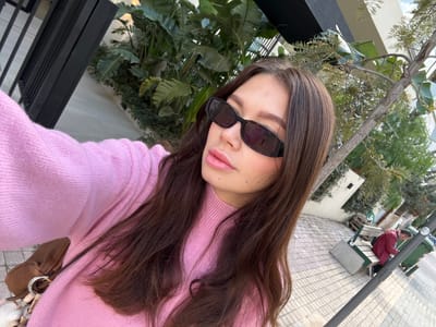 The scene depicts an outdoor urban environment in the Municipal Unit of Elliniko, Greece, captured from a first-person perspective, likely a selfie. A female subject occupies the foreground and center-left, presenting her upper body and face. She has brown hair and wears a light pink long-sleeved sweater. Black, rectangular-framed sunglasses with "GUCCI" branding on the left temple obscure her eyes. Her lips are visible and coated with pink lip gloss. A brown bag with a silver-colored ring attachment hangs from her shoulder, partially visible on her left side.
To the left of the subject, a dark metal fence or railing with vertical bars is present, behind which dense green foliage, including large-leafed plants and smaller shrubs, borders a light-colored building. To the subject's right, a paved area with light-colored rectangular pavers is visible, extending into the background. A tree with a thin trunk and sparse green foliage is positioned in the mid-ground. Further back, a dark gray street light pole stands. A green public bench with wooden slats and light gray metal supports is situated to the right, on which a male individual, wearing a red jacket and dark pants, is seated, facing away from the camera. A green refuse bin is located behind the bench. The scene is under bright natural daylight.
