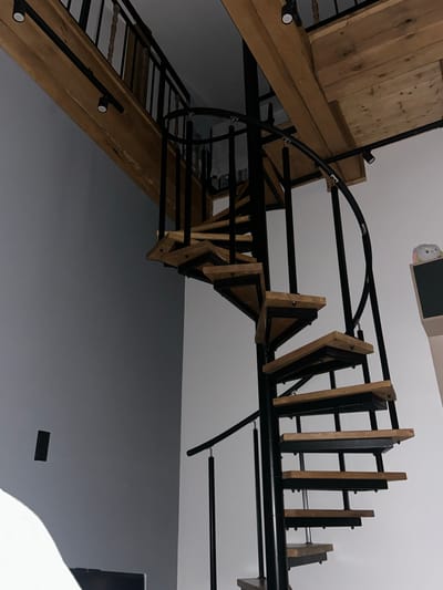 The image shows an interior view of a modern spiral staircase with wooden treads and a black metal frame. The staircase ascends upwards and curves to the right, with a black metal railing following its path. Above the staircase, a wooden beam with two track lights runs horizontally. The walls appear to be painted in two different colors: a dark gray on the left and a lighter off-white on the right. A black rectangular electrical outlet is visible on the dark gray wall. On the off-white wall, a shelf unit with a black display area and a small, plush toy resembling a white bunny with pink accents is placed on top. The overall scene suggests a residential interior, possibly a loft or a home with multiple levels. The location in Siedlce, Poland is not discernable from the image itself.