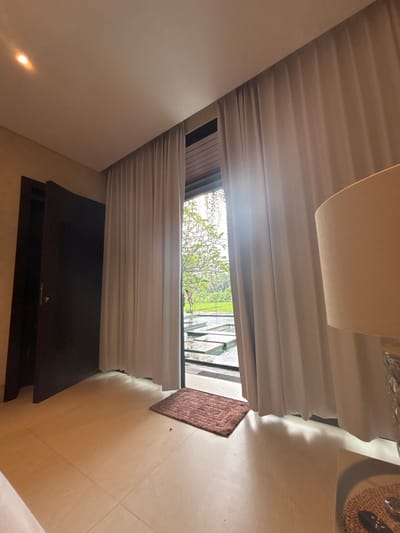 A room with light beige walls and tile flooring featuring a closed dark wood cabinet against one wall. A window is framed by white curtains, offering a view of greenery. A brown rug sits on the floor near the window. The top of a lamp is visible in the upper center of the image. The photo appears to be taken at an angled perspective, making the room appear tilted.