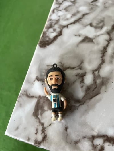 A small, detailed figurine of a soccer player, likely representing Lionel Messi, is positioned on a marble-patterned surface. The figurine is wearing an Argentina national team jersey with the number 10. Its right arm is raised in a waving gesture. The background consists of a blurred green surface. There are no discernible elements in the image that can be linked to the city of Vărzărești, Moldova.
