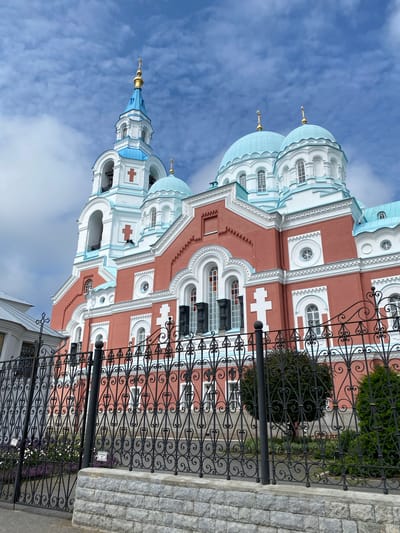 The image shows the exterior of a grand, pink and white Orthodox church with blue domes, located in Sортавала, Russia. A decorative black metal fence with a stone base stands in the foreground, partially obscuring the view of the church's lower sections. The church features multiple onion domes topped with golden crosses, a bell tower with arches and red crosses, and a facade adorned with white decorative elements and arched windows. The sky is blue with scattered white clouds. No people are visible in the image.