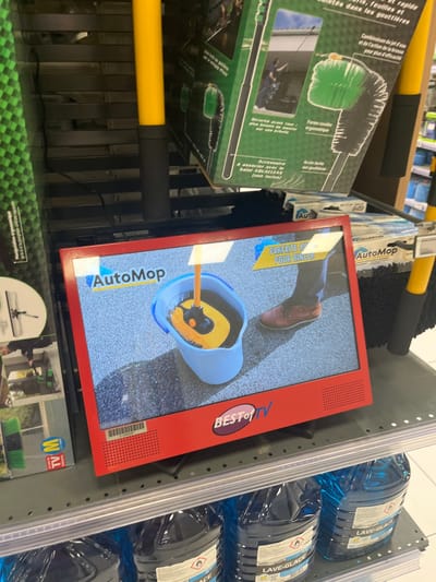 The image shows a red, small TV screen playing a commercial for an Auto Mop on a shelf in a retail store located in Saint-Brice-Courcelles, France (latitude: 49.283, longitude: 4). The TV screen shows a person's foot in brown shoes stepping on a gray, textured surface next to a blue bucket filled with soapy water and a yellow mop handle. The mop head is partially submerged in the water, and a yellow mop handle is visible. The TV screen is tilted forward and slightly to the right. The screen is on a black shelf with white dots. Above the TV screen are product boxes featuring pictures of cleaning tools and accessories. Three blue containers of glass cleaner are visible below the shelf. The container labels say "LAVE-GLACE". One container, partially visible, says "ACE".