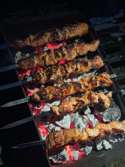 The image shows six metal skewers holding pieces of grilled meat, arranged over a grill filled with glowing red coals and ash. The meat appears cooked with some charring. The skewers are placed on the edges of the grill. Some of the charcoal appears burnt and covered with white ash. The setting seems to be outdoors, based on the darker environment.
There is no evidence to support that the location is in the city of Verkh-Katunskoye, Russia. The specific location cannot be determined from the image.
No people are visible.