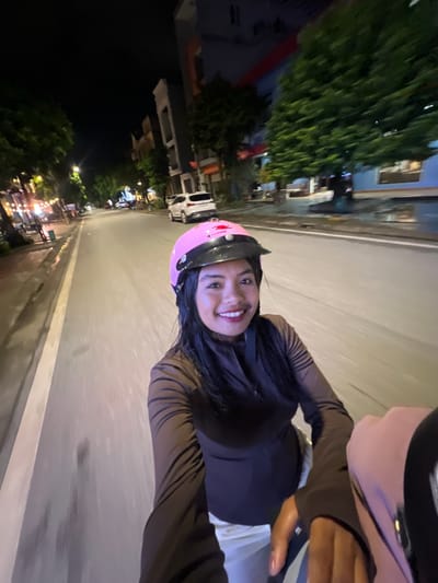 A woman with long dark hair, wearing a pink helmet and a brown long-sleeved shirt, smiles at the camera while riding on a motorcycle at night on a paved road in Minh Xuân Ward, Vietnam. She is looking towards the camera, and her face is well-lit. The road is wet, indicating recent rain, and there are streetlights and buildings lining the street on either side. A white SUV is visible in the background on the right side of the road. The image appears to have been taken from the perspective of someone riding on the back of the motorcycle.