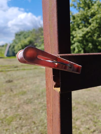 A close-up, eye-level shot focuses on a single, pink plastic clothespin attached to a rusty metal post. The clothespin is positioned horizontally, extending from the left to the right of the frame, and appears to be holding something unseen. The metal post, a vertical element, is weathered and shows significant signs of rust. Behind the post and clothespin, the background is blurred, depicting an outdoor scene under a bright blue sky with scattered white clouds. The ground appears to be dry grass or dirt, with some greenery and trees visible in the distance. A building with a slanted roof is also faintly visible in the far background, to the left. The image is likely taken in an outdoor, possibly rural, setting in the village of Shiryaevo, Russia.