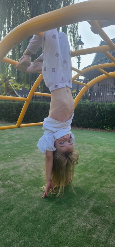 A young girl with blonde hair and blue eyes is upside down on a yellow jungle gym in a park, likely in Groenlo, Netherlands. She is wearing a white t-shirt and white pants with a pattern. Her arms are outstretched, and her hands are touching the green, artificial turf ground. The background shows trees, a street lamp, and a building. The lighting suggests it is daytime, with the sun casting shadows.