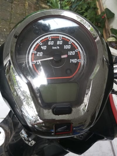 A close-up shot shows a motorcycle's speedometer. The dial is black with white numbers indicating kilometers per hour, ranging from 0 to 140. A red-orange accent line highlights the outer edge of the speed indicators. The needle points near the 20 km/h mark. Below the speedometer is a digital display screen and a black button. The surrounding chrome casing reflects light, showing environmental elements. A Honda logo is visible at the bottom.