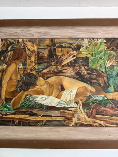 An oil painting depicts two nude figures in a rustic, outdoor setting. The figure in the foreground is lying on their side, propped up by their elbow, with a white cloth draped over their lower body. A second figure is seated to the left, facing away from the viewer, holding a white flower. The background is a mix of natural elements, including wood, foliage, and what appears to be water. The painting is framed in wood and appears to be hung on a wall, with the top of the frame against a white surface. The painting is signed in the lower right corner. The scene is not specific to Sunny Isles Beach, United States of America, and no details indicate a particular city or location.