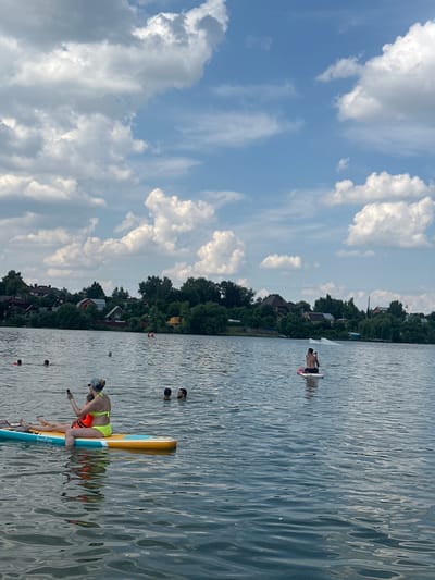 The image depicts a body of water, likely a lake or river, on a cloudy day. In the foreground, two people are on a yellow and blue paddleboard. One person is seated and appears to be holding a phone, while the other is lying down. Further out on the water, a person is standing on a paddleboard. Several other individuals are visible swimming in the water. In the background, a tree-lined shore with houses and buildings is present. The sky is filled with dramatic white and gray clouds against a blue sky. The overall scene suggests a recreational activity taking place in an outdoor setting. The location is identified as Dal'nie Prudishchi, Russia.