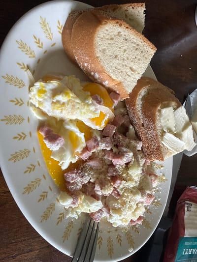 A plate of food sits on a dark wooden table. The plate is white with a pattern of golden leaves around the rim. On the plate are two slices of bread, one topped with several slices of butter. Next to the bread are scrambled eggs with diced ham and grated cheese. A fork rests on the side of the plate, its tines in the food. In the background, a package of cheese is visible. The lighting suggests it is daytime.