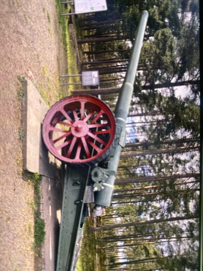 The image shows a large, green artillery piece positioned outdoors on a concrete slab, with a prominent red-spoked wheel. The artillery piece is angled diagonally, with its barrel extending upwards and towards the right. The background consists of a dense forest of tall, thin trees, with dappled sunlight filtering through the canopy. A gravel path with some sparse grass and yellow flowers is visible to the left of the concrete slab. A sign or informational board is partially visible in the upper right corner. This scene is located in Kerava, Finland.