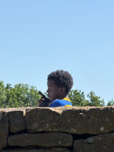 A low-angle, medium shot shows a dark-skinned young man peering into a black instrument. He is positioned behind a rough-textured stone wall that angles vertically. The man has short, dark curly hair, and he is wearing a blue shirt with a yellow shoulder stripe. The backdrop includes a clear blue sky and the foliage of green trees. The location is the city of Fentange, Luxembourg.