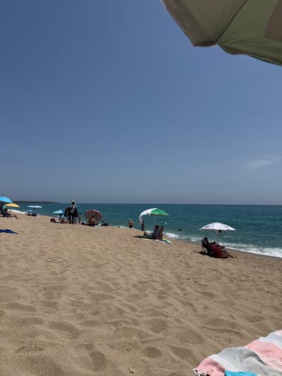 The image depicts a crowded beach scene on a sunny day. The foreground is dominated by sand, with some beach towels and debris scattered on it. In the middle ground, several beach umbrellas provide shade for sunbathers. A person is seen standing near some umbrellas, possibly a vendor. People are scattered across the sand, some sitting, some standing, and some are in the shallow water. The sea is a vibrant blue with gentle waves. The horizon is visible, with a clear blue sky above. A small island or peninsula is visible in the distance on the left. The scene suggests a typical summer day at a popular beach resort.