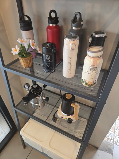 The image displays a multi-tiered metal shelf unit, likely located indoors, possibly in Klapanunggal, Indonesia, given the context. The top shelf holds several water bottles of various colors and designs, along with a small potted artificial plant with white flowers. The bottles are arranged in a row, with a beige bottle with a black lid, a red bottle with a black lid, a dark gray bottle with a black lid, a white bottle with a light pink lid and a decorative drawing of coffee beans, and another white bottle with a beige lid and a scenic illustration.

The middle shelf contains a vintage-style coffee maker with a pressure gauge and a black kettle with a wooden handle sitting on a small electric heating element. The kettle is turned off.

The bottom shelf of the unit is partially visible and holds a cream-colored storage container with a lid. The overall environment appears to be a clean, organized space, possibly a home or a retail display. The lighting suggests an indoor setting with natural light filtering in from the side. There are no people or animals present in the image.