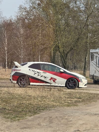 The scene, located in Wittenberg, Germany, presents a white Honda Civic Type R (FN2 generation) parked on an unpaved, uneven ground surface composed of dry soil and sparse, light-colored vegetation. The vehicle is oriented in profile, facing right, displaying a distinctive livery featuring red and black graphic elements, including the text "CIVIC TYPE R" and a prominent "R" logo on its side. It is equipped with dark multi-spoke alloy wheels and a rear spoiler is visible. In the background, a line of deciduous trees stands, characterized by bare branches and light brown foliage, indicative of late autumn, winter, or early spring conditions. Some trees exhibit light-colored bark, while others are darker with visible lichen or moss on their trunks and branches. To the far right of the frame, the front section of a white recreational vehicle (RV) or camper, featuring a dark vertical stripe and window, is partially visible behind the Civic. No individuals are present in the image.