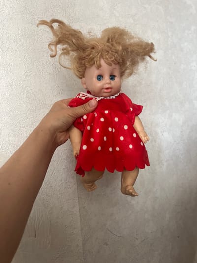 A person's hand is holding a vintage doll with blonde, messy hair and blue eyes, wearing a red polka dot dress. The doll is suspended against a textured, off-white wall.