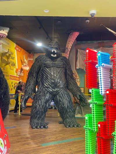 The image shows a life-size statue of a gorilla in a museum-like setting. The gorilla is dark gray and appears to be made of textured material, possibly resembling tire rubber. It is positioned in a dynamic pose, with its arms and legs spread.

In the background, there are colorful, hanging decorations that look like upside-down accordion-like structures in red, blue, white, and green. There are also displays with text and images, including a shark jaw and a kangaroo. A person wearing a green shirt and black pants is visible standing near the bottom right of the image, appearing to interact with one of the displays or the gorilla.

Visible text includes "SURF'S UP!", "GREAT SHARK JAW", "RARE ALBINO KANGAROO", and "UNBELIEVABLE". The overall scene suggests an indoor exhibition, possibly at an amusement park or a museum with a focus on natural history or curiosities. The lighting is artificial, indicating it's likely daytime or evening indoors. The presence of the text "SURF'S UP!" and "Surfers Paradise, Australia" in the prompt might suggest a connection to the location, though the image itself doesn't explicitly confirm this. The vibe is playful and educational, aimed at engaging visitors.