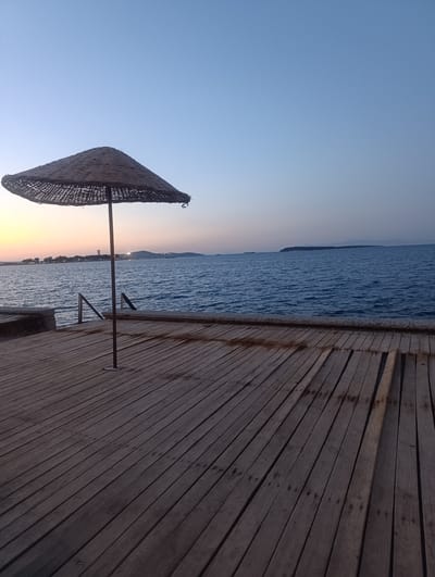 This image captures a serene seaside scene at dusk in Urla, Turkey. The foreground is dominated by a wooden deck, its planks stretching diagonally from the bottom left to the top right of the frame, showing signs of wear and tear. A single thatched umbrella stands upright on the deck, its dark, woven material contrasting with the lighter wood. Beyond the deck, a calm, blue sea stretches out to the horizon.

In the distance, a coastline is visible, dotted with buildings and trees, hinting at a town or settlement. A tall, slender structure, possibly a lighthouse or a tower, stands out against the sky on the left side of the distant landmass. The sky transitions from a soft yellow and orange glow near the horizon, suggesting the last rays of the setting sun, to a clear, pale blue at the top of the image. The overall mood is peaceful and tranquil, evoking a sense of quiet end-of-day relaxation. The time of day is clearly sunset or twilight, with the soft lighting and clear sky indicating fair weather. There is no visible text in the image.
