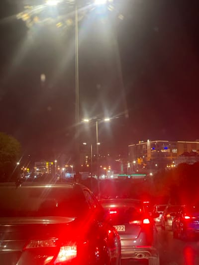 The image presents a nighttime urban street scene in Dabouq, Jordan, viewed from within a vehicle. In the immediate foreground, multiple vehicles are visible, displaying illuminated red brake lights, indicative of stopped or slow-moving traffic. A dark-colored sedan occupies the left foreground, its taillights brightly illuminated. Directly ahead, a silver/grey sedan shows a partially visible white license plate with the numerical sequence "9272".
The upper portion of the scene is dominated by several intense artificial light sources. A very tall mast-mounted light fixture, equipped with multiple luminaires, emits powerful light causing significant lens flare and starburst effects. Below this, standard streetlights are also visible, contributing to the overall illumination.
In the background, an urban landscape unfolds, featuring numerous illuminated multi-story buildings on an elevated terrain. A prominent building on the right exhibits distinctive orange and blue architectural lighting, complemented by yellow decorative lights. Other commercial structures and signs are discernible through the ambient light. The sky is dark, consistent with nighttime conditions. No individuals are explicitly visible within the frame.