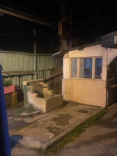 This image captures a dimly lit outdoor scene, possibly in Surami, Georgia, at night. The foreground features a weathered concrete pathway with patches of grass growing through cracks. To the right, a simple, light-colored wooden structure with several windows stands, resembling a small kiosk or shed. In the center, a low, concrete structure with steps, possibly a decorative fountain or water feature, is visible. A single faucet is attached to the structure. To the left, a green wooden fence creates a backdrop. The overall impression is one of a somewhat neglected, utilitarian outdoor space.