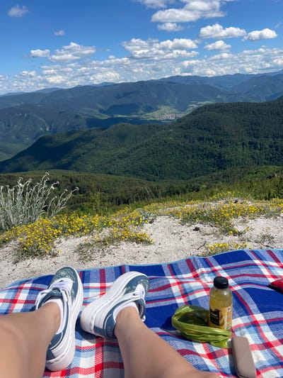 The image depicts a person's legs and feet wearing white and blue sneakers, resting on a red, white, and blue checkered blanket. To the right of the legs are a bottle of yellow liquid and a green bag. The blanket is laid out on a rocky, sandy surface covered with small yellow flowers. In the background, a lush green mountainous landscape unfolds under a bright blue sky dotted with white clouds. The location is identified as Kosovo, Bulgaria, suggesting an outdoor recreational setting.