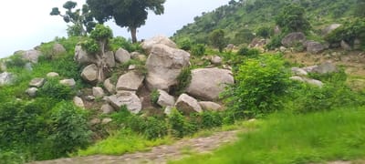 The image depicts a rocky, hilly landscape covered in lush green vegetation. Large boulders are scattered across the slopes, interspersed with trees and dense bushes. A path, partially visible, winds through the scene. The environment appears natural and undeveloped, likely an outdoor setting outside of an urban area. The image was taken from a moving vehicle, as evidenced by the motion blur in the foreground, suggesting a scenic drive through the countryside. The lighting suggests an overcast day. No people or specific structures are visible. This landscape is characteristic of areas surrounding Gu, Nigeria, known for its hilly terrain and natural beauty.