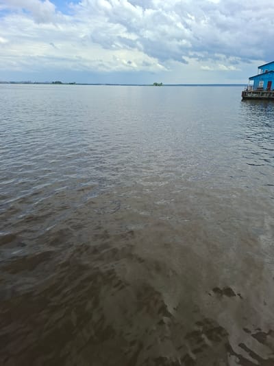 This is a landscape photograph taken from a dock or pier, featuring a view of a large body of water under a cloudy sky. On the left side of the image, a section of a bright blue building with red accents is visible, suggesting it is a structure on the shore or a boathouse. The building has windows and a railing, indicating it's accessible.
The majority of the frame is dominated by the water, which appears somewhat murky or silted, with ripples and light reflections creating texture across its surface. The water extends to the horizon, where it meets a hazy outline of distant land, possibly featuring trees and some structures, characteristic of a shoreline. The sky is filled with dramatic, white and grey clouds, with patches of blue sky peeking through, suggesting either an overcast day with breaks of sun or a time of day when the clouds are particularly active.
The scene evokes a sense of calm and perhaps a touch of melancholy due to the cloudy sky. The location context of Verhnij Uslon, Russia, implies a riverside or lakeside setting, common for such structures. The time of day is not explicitly clear but likely daytime due to the brightness of the sky and the reflections on the water. There is no visible text in the image.