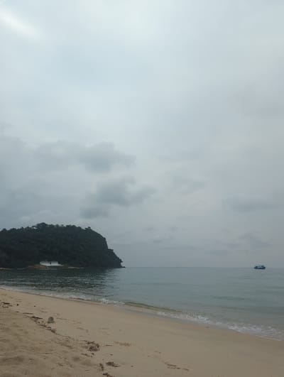 A coastal scene under a cloudy sky. The right side shows a sandy beach with gentle waves touching the shore. The water transitions from light to darker shades. A green, hilly land mass juts out into the water near the center. A distant boat can be seen on the water's surface towards the upper right. The sky takes up the majority of the image, filled with gray clouds.