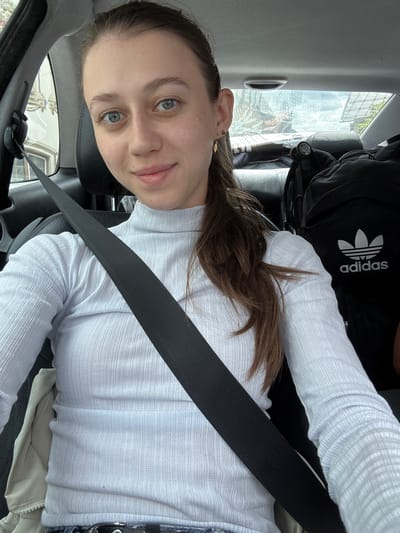 In the city of Reutte, Austria, a young Caucasian woman with light brown hair, blue eyes, and fair skin is photographed from the chest up inside a car. She is wearing a white, long-sleeved, ribbed turtleneck sweater and a black seatbelt is fastened across her chest. She is looking directly at the camera with a slight smile. In the background, a black backpack with the Adidas logo is visible on the car seat behind her. The car's interior, including the headrest and ceiling, is also visible.