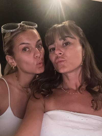 Two women are taking a selfie. The woman on the left has blonde hair pulled back and is wearing a white tank top and a necklace. She has sunglasses on top of her head. The woman on the right has brown hair and is wearing a strapless white top and a beaded necklace. Both women are puckering their lips. The background is dark, and there is a bright light source from above and to the left. The location is specified as Savigneux, France.