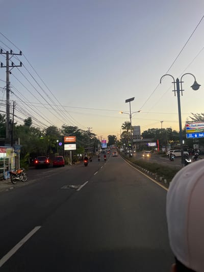 The image depicts a street scene in Manado, Indonesia, likely during twilight or early evening. A road curves to the right, with a lane of traffic moving away from the viewer. Several motorcycles are visible on the road, along with a few cars, suggesting moderate traffic.
On the left side of the road, there are numerous utility poles with a dense network of electrical wires. Beyond the poles, a line of trees and vegetation can be seen. Several illuminated billboards and signs are interspersed along the roadside. One prominent sign reads "Indomaret" and "Fresh" in bold lettering, indicating a convenience store. Another sign shows "Klausei Bar" and "Salebo Bagindo". Further down the road, more signs and buildings are visible, some with neon lights.
The sky is a pale gradient of blue and orange, characteristic of a sunset or sunrise. The lighting suggests the transition from day to night. A portion of a person's white headwear is visible at the top right of the frame, implying the photo was taken by someone moving along the street, possibly on a motorcycle or in a vehicle. The overall atmosphere is that of a busy urban or suburban street at dusk.